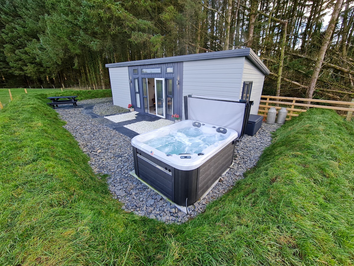 A modern lodge is set amidst greenery, accompanied by a hot tub featuring bubbling water. The exterior is accessible, with a pebble pathway leading to a picnic area. Tall trees provide a natural backdrop, enhancing the secluded atmosphere.