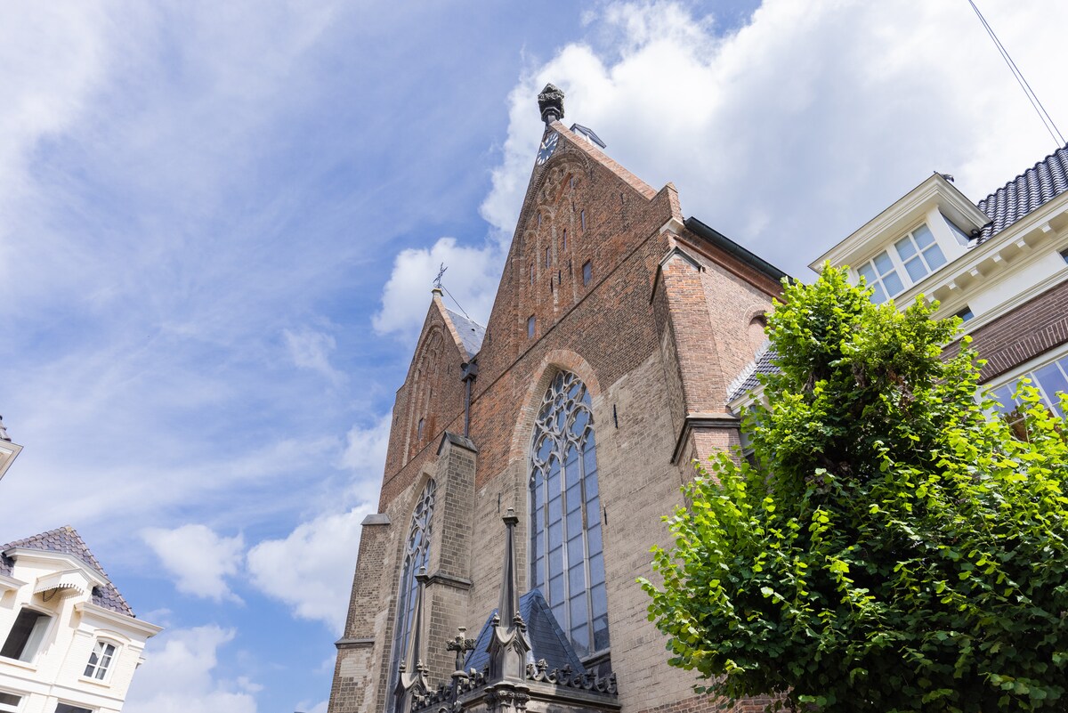 A majestic façade of the Broederenkerk is visible, showcasing intricate brickwork and large stained glass windows. The structure features pointed arches and a tower, framed by lush greenery under a bright blue sky with scattered clouds.