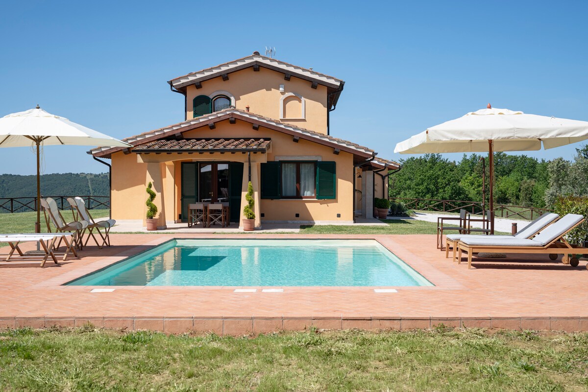 A spacious villa is depicted, featuring a welcoming exterior with a tiled roof and balconies. A private pool is surrounded by sun-loungers and umbrellas, set against a backdrop of green hills and olive trees. Clear blue skies enhance the scenery.