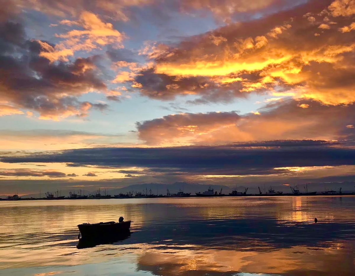 A tranquil seascape is depicted, featuring gentle waves reflecting vibrant orange and purple hues from the sky at sunset. A small boat floats calmly in the foreground, while silhouettes of fishing boats are visible in the distance against a backdrop of clouds.