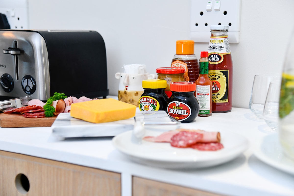 A countertop displays various food items, including slices of cheese, meats, and condiments in jars and bottles. A toaster is positioned to the left, and a plate with meat slices is placed in the foreground. Additional items, such as marmite and mustard, add color to the arrangement.