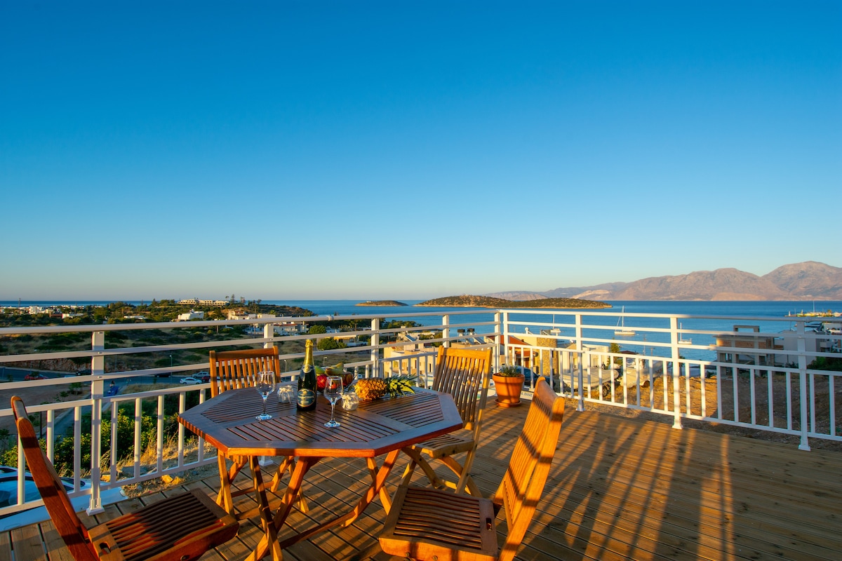 A spacious outdoor terrace features a wooden dining table surrounded by four chairs, offering a scenic view of the bay. The clear blue sky and distant mountains frame the horizon, enhancing the tranquil atmosphere of the space.