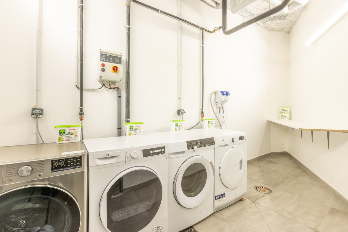 The laundry area features a modern washing machine and dryer, both available for guest use. The space is clean and well-lit, with a simple countertop against the wall for folding clothes. Laundry detergent and supplies are organized on shelves above the appliances.
