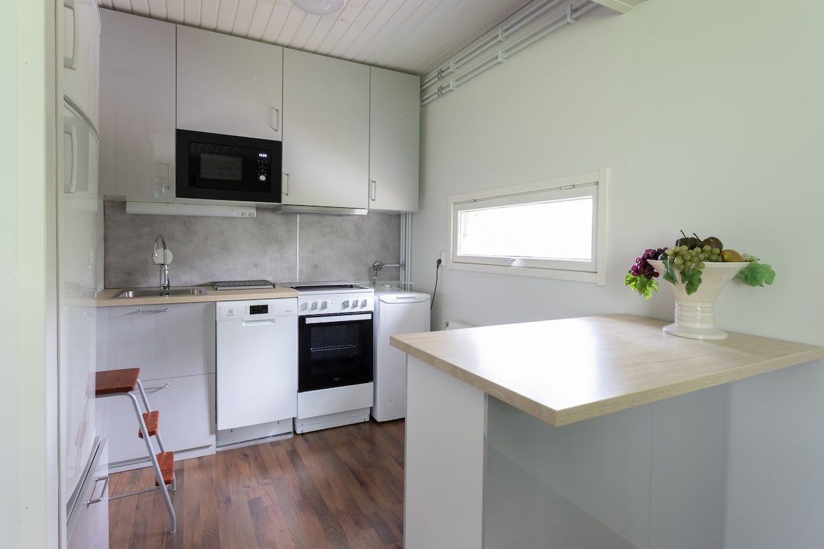 A functional kitchen area is highlighted by modern appliances, including a stove and microwave. Natural light filters through a nearby window, illuminating the light-colored cabinetry. A simple dining space is created by a countertop, and a small step stool is positioned nearby.