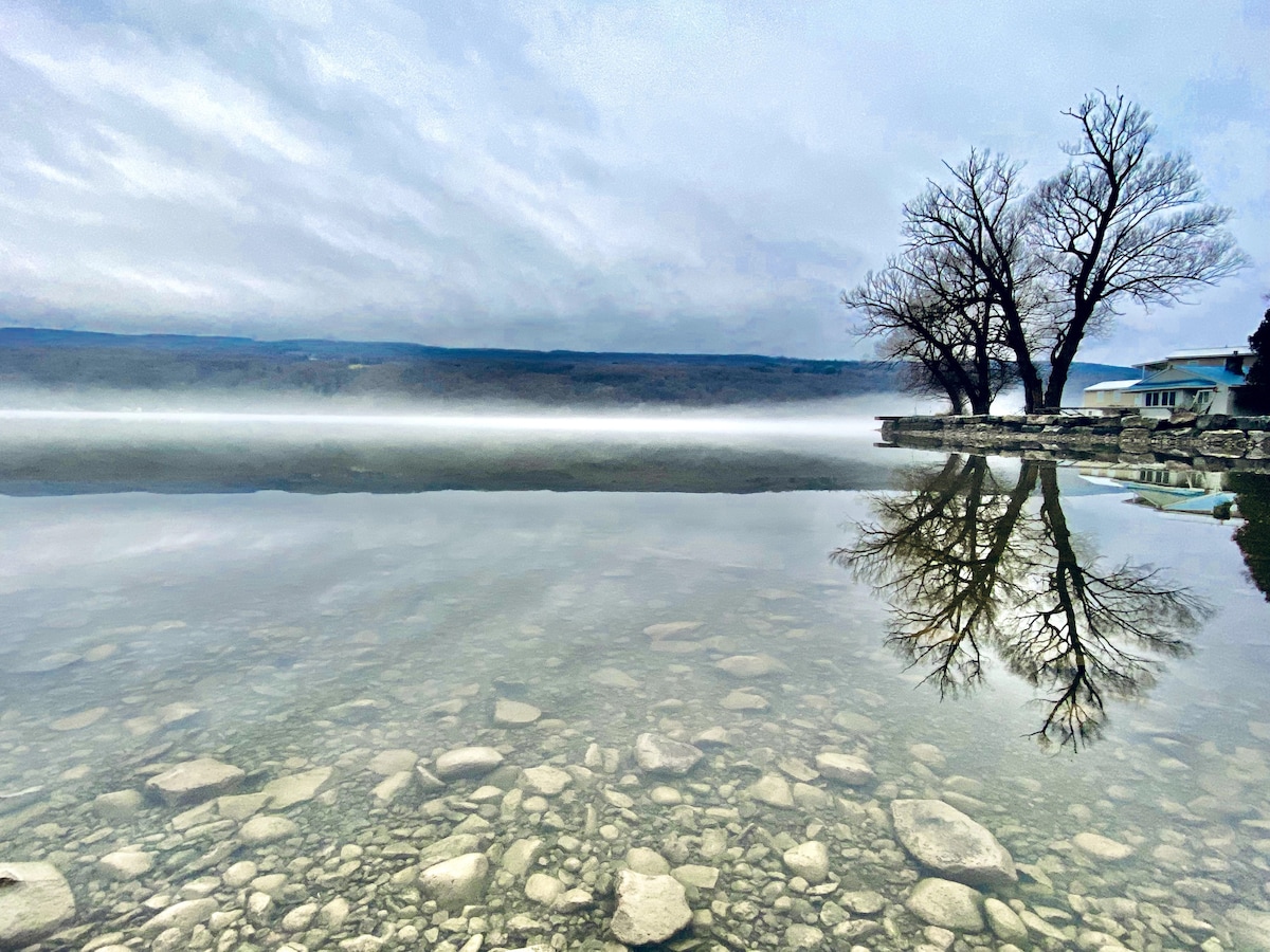 A serene lakeside view captures still water reflecting the overcast sky and nearby trees. The shoreline is bordered by smooth stones, while a soft mist rises over the surface of the lake, creating a tranquil atmosphere.