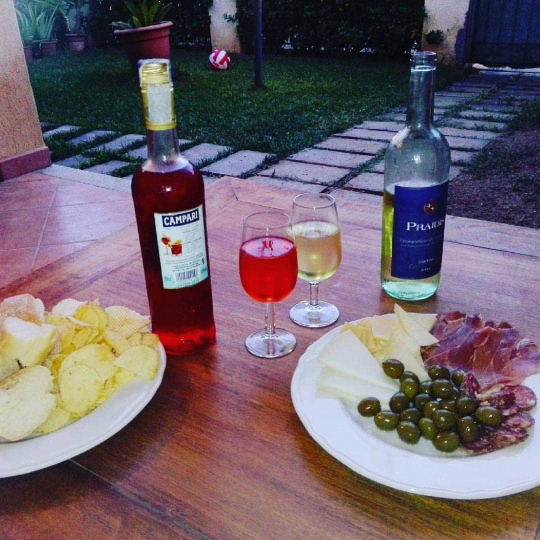 A wooden table is set outdoors, featuring an assortment of snacks. Plates contain crispy chips, olives, cheese, and sliced cured meat. Two glasses filled with brightly colored beverages are positioned beside the food, with a bottle of wine placed on the table. A grassy garden is visible in the background.