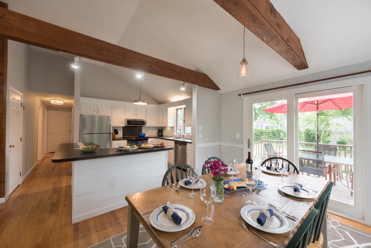 A spacious dining area features a wooden table set for four, complemented by dark green chairs. Natural light streams in through glass doors leading to a deck, where an umbrella provides shade. The kitchen area is visible in the background, with modern appliances and ample counter space.