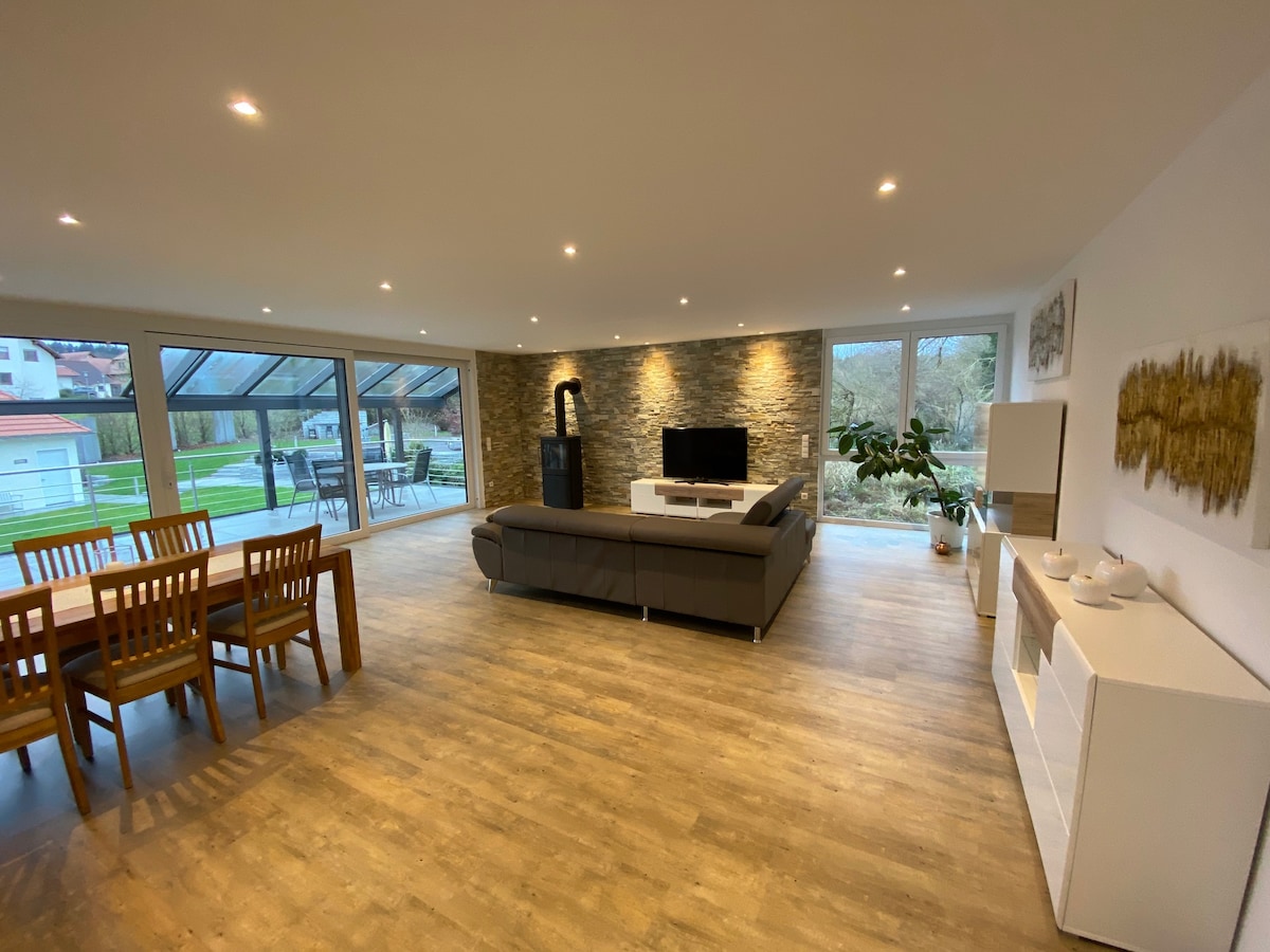 A spacious living and dining area features a wooden dining table with six chairs. A comfortable gray sectional sofa is positioned towards a flat-screen TV and a modern wood-burning stove. Large windows provide ample natural light and overlook the outdoor space.