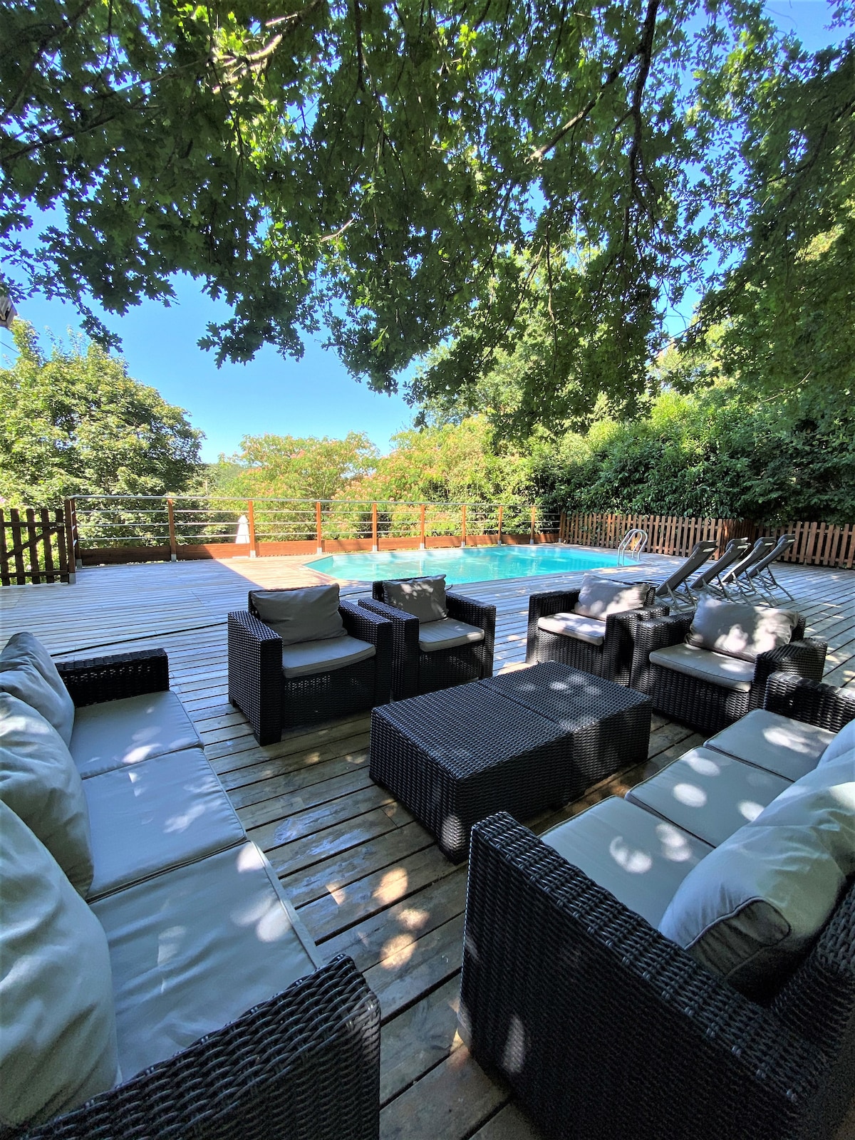 An outdoor seating area features comfortable wicker furniture set upon a wooden deck. The space is surrounded by lush greenery and overlooks a private swimming pool. Sun loungers are positioned near the pool, inviting relaxation under a clear blue sky.