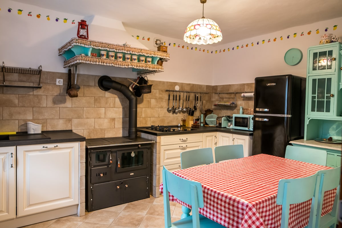 The kitchen features a vintage design with a patterned red-and-white tablecloth covering a wooden dining table. A classic black stove and modern appliances, including a turquoise refrigerator and microwave, complement the beige tiled walls. Decorative accents, such as a colorful overhead light, are visible.