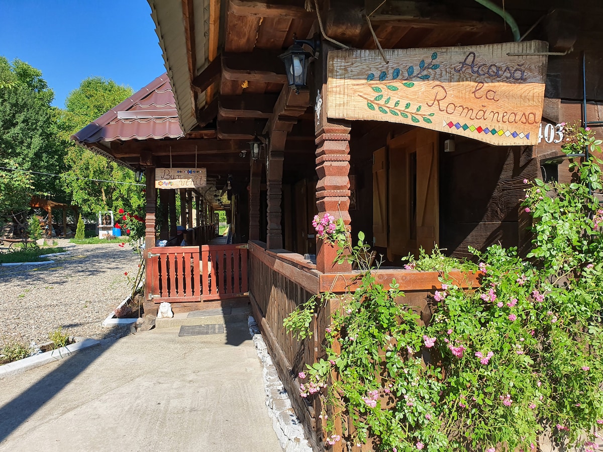 The entrance of a traditional wooden house features carved details and signage that reads 'Acasa la Romaneasa'. Vibrant flowers adorn the front porch, complemented by a gravel pathway lined with greenery. The inviting porch area is ideal for relaxation.
