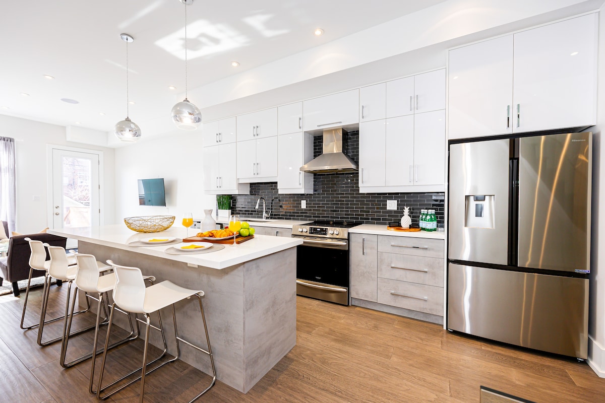 A modern kitchen features sleek cabinetry in white and gray tones. Stainless steel appliances, including a refrigerator and oven, are present. An island with seating is complemented by a stylish bowl of fruit, and large windows allow for natural light to brighten the space.
