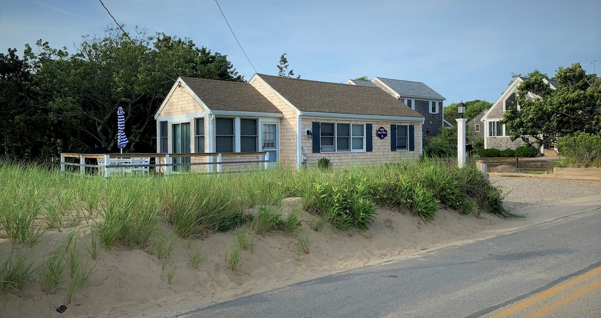 A charming cottage is seen from a distance, surrounded by lush green grass and decorative fencing. Soft siding and a sloped roof complement the coastal environment. A nearby road and sandy path lead towards the beach, enhancing the cottage's proximity to the shore.