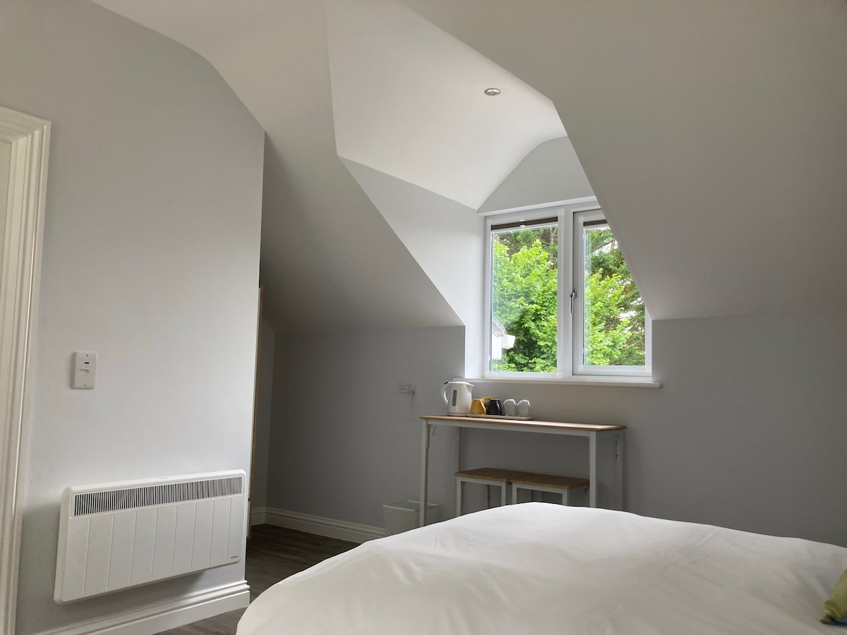 A bright room showcases an angled ceiling and large windows allowing natural light to fill the space. A small table with tea-making facilities is positioned beside the window. A spacious bed is located in the foreground, inviting relaxation.
