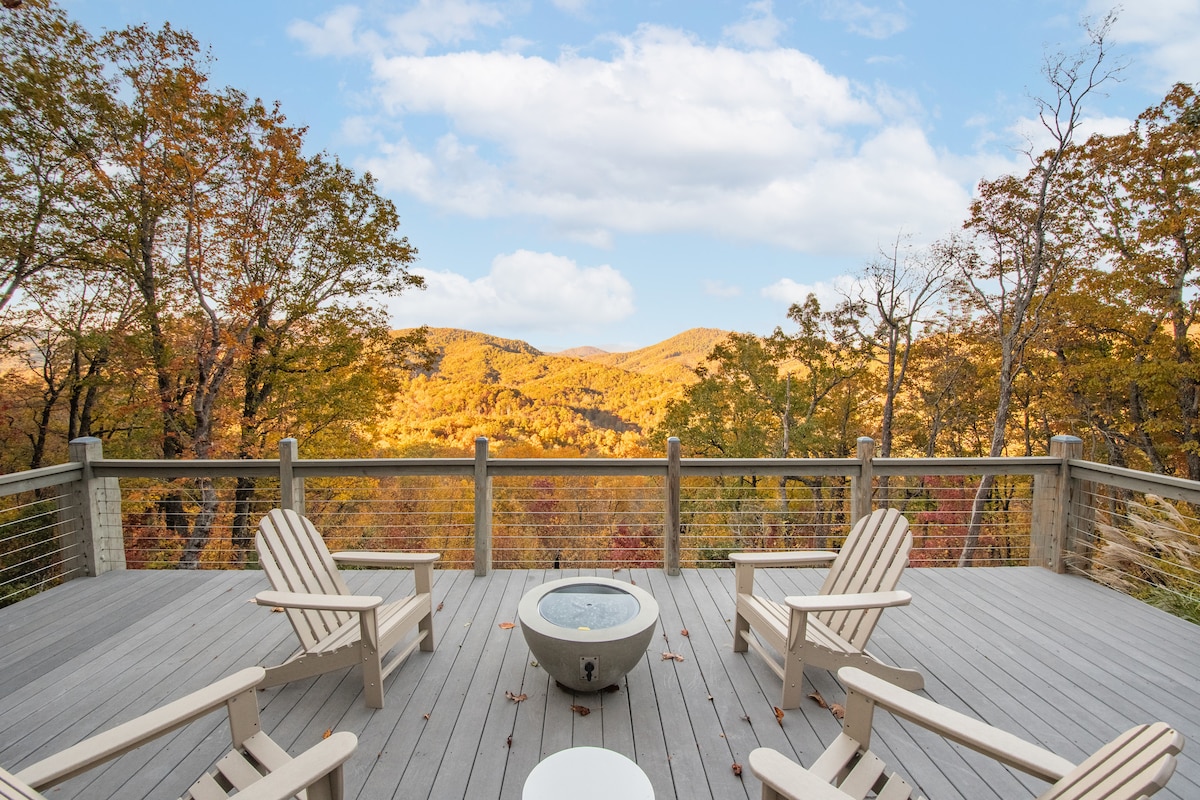 A spacious deck showcases several Adirondack chairs arranged around a fire pit, with panoramic views of colorful autumn foliage across the mountains. The landscape features rolling hills and a bright sky, creating a serene outdoor space for relaxation.
