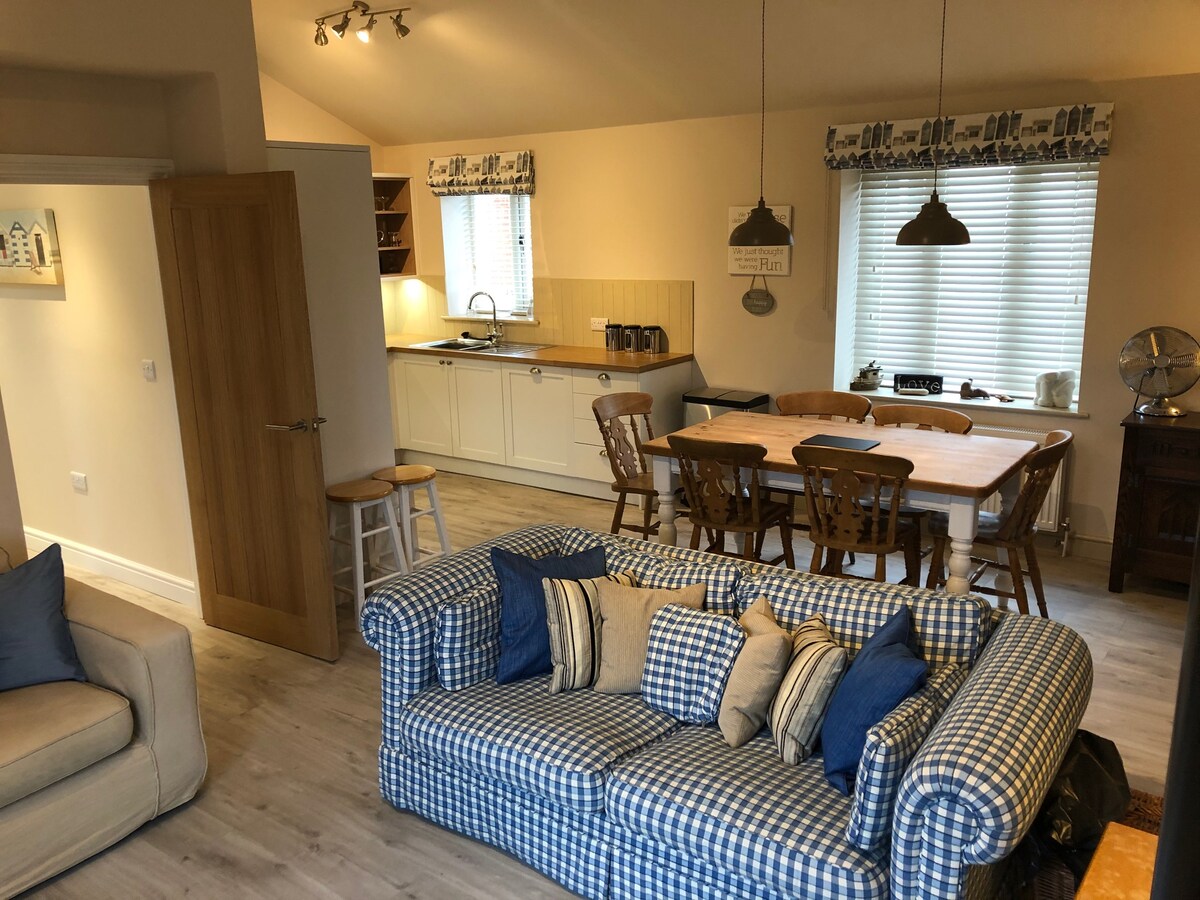 A cozy living space features a checked sofa adorned with cushions, complemented by a dining area with wooden chairs and table. The adjacent kitchen area includes cabinets and countertop appliances. Natural light streams through the window treatments, enhancing the welcoming ambiance of the room.