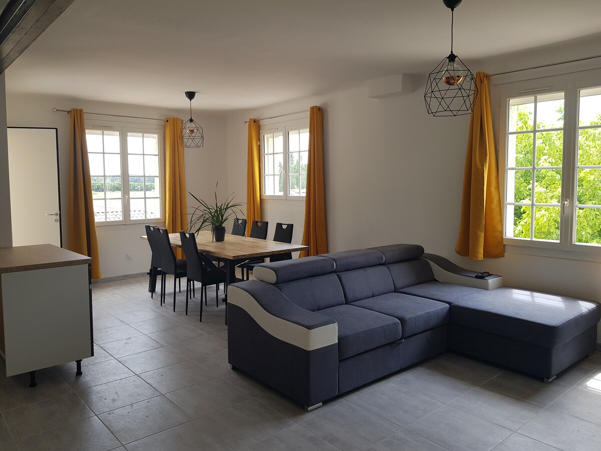 The spacious living area features a modern sectional sofa positioned near large windows, allowing natural light to fill the room. A wooden dining table is surrounded by six chairs, with yellow curtains adding a bright touch to the neutral decor.