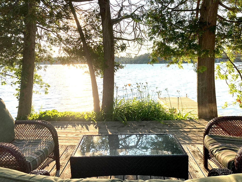 A lakeside deck is framed by trees, showcasing a view of the water reflecting the sunset. Two wicker chairs with cushions and a glass-top table are positioned for relaxation, while greenery and wildflowers add to the natural setting.