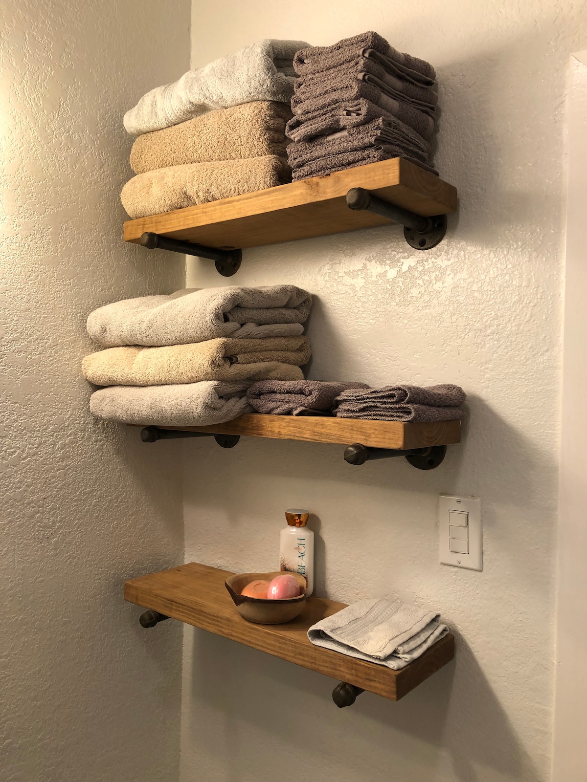 Two wooden shelves are mounted on a bathroom wall, showcasing neatly stacked towels in shades of beige and gray. A small tray holds a soap dispenser and a decorative item on the lower shelf. A soft glow from a nearby light fixture enhances the tidy arrangement.
