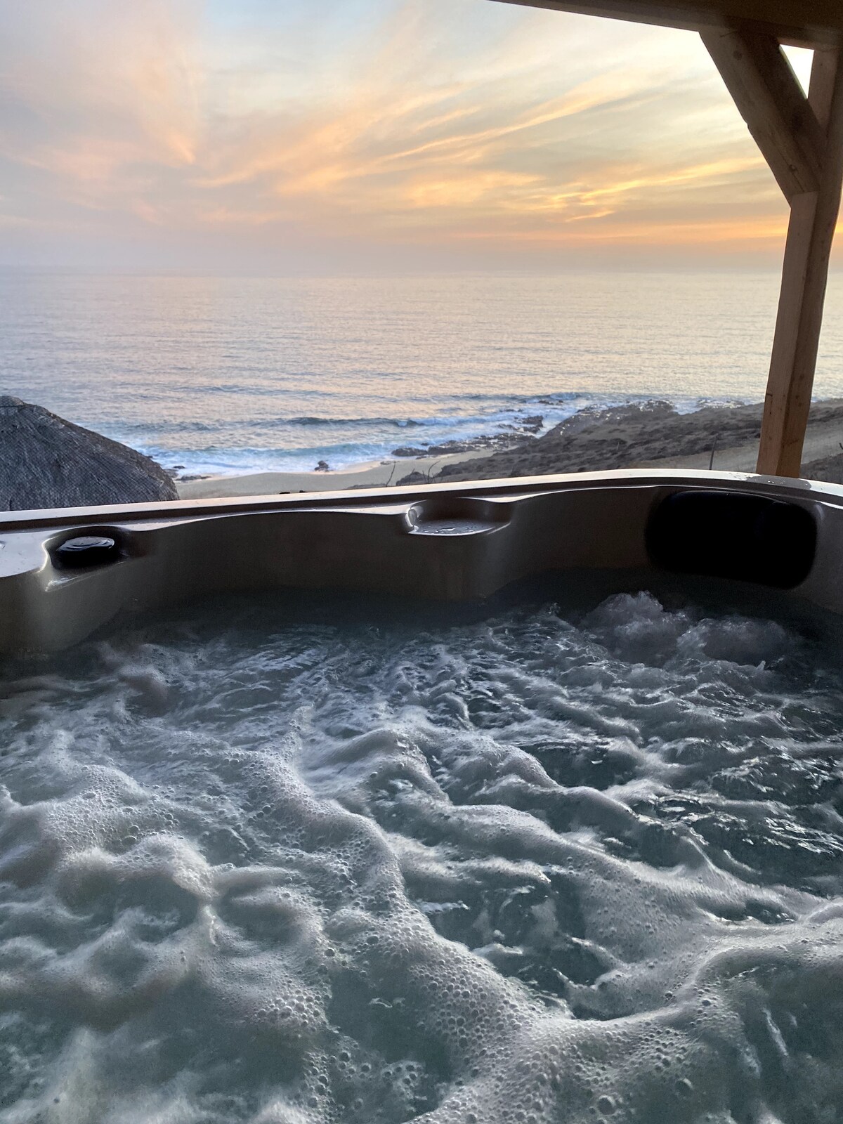 A hot tub sits beneath a covered area, offering views of the ocean waves gently rolling against the rocky shore. The sky features soft pastel colors as the sun sets on the horizon, creating a peaceful and serene atmosphere.