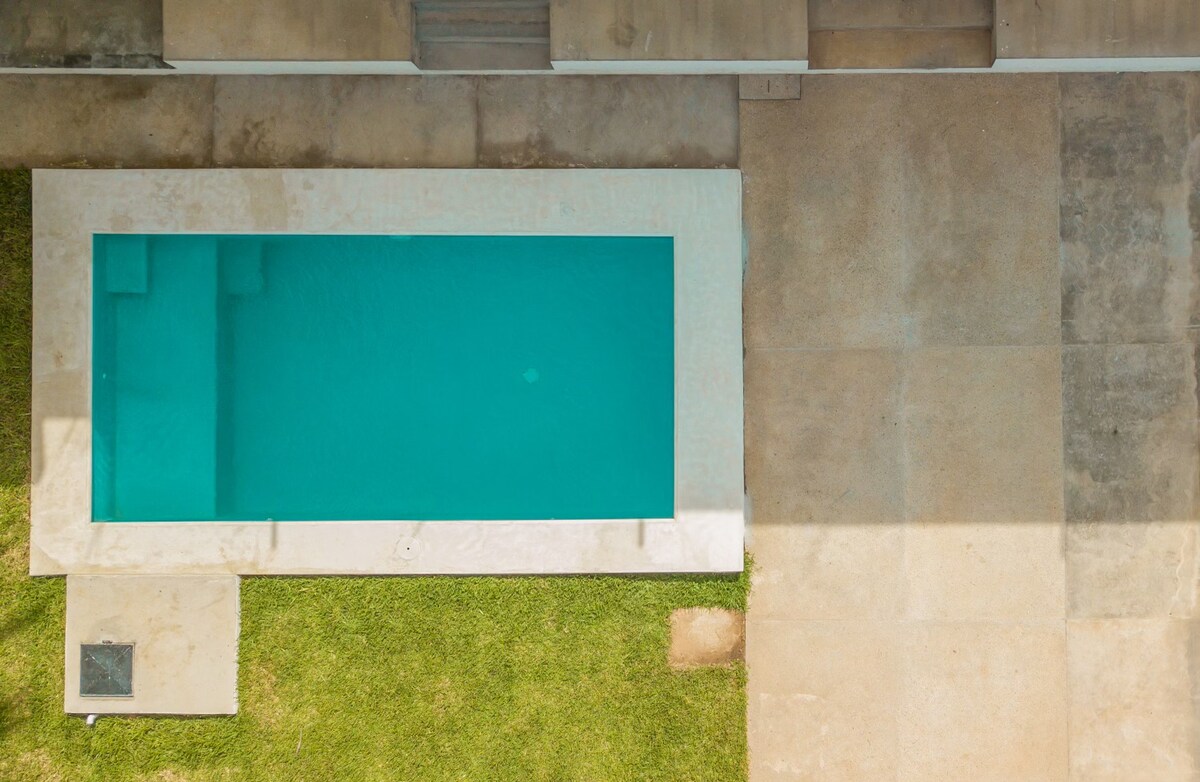 An aerial view showcases a rectangular swimming pool surrounded by a smooth concrete patio. Green grass borders the pool, providing a natural contrast to the cool blue water. The pool area appears well-maintained, offering ample space for relaxation.