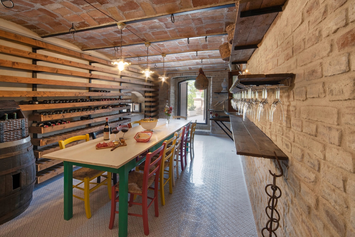 A rustic dining area is characterized by exposed brick walls and wooden shelves lined with various wine bottles. A long table surrounded by colorful chairs invites gatherings, while glassware hangs from a wooden rack. Soft lighting from pendant fixtures enhances the cozy atmosphere.