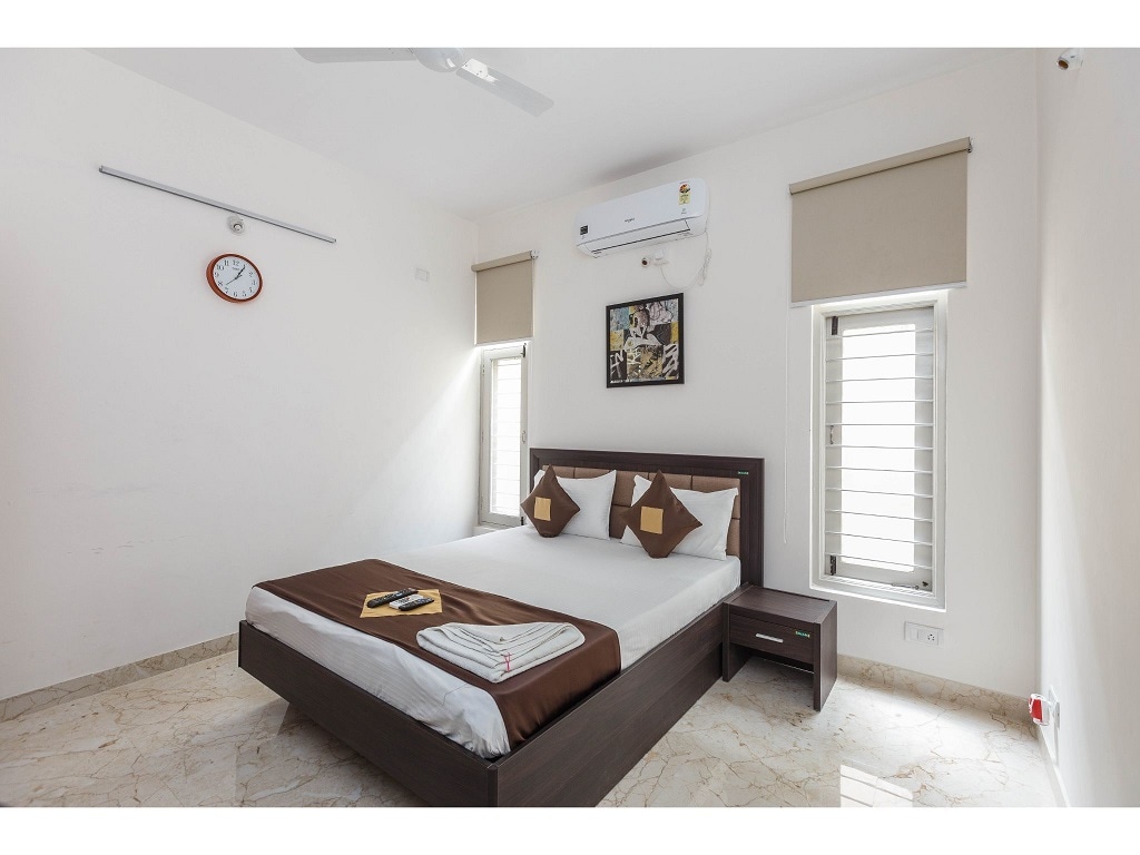 A well-lit bedroom features a spacious bed with a dark brown bedspread and two accent pillows. A bedside table holds a clock, while windows allow natural light to fill the room. Wall art adds a touch of character to the neutral decor.