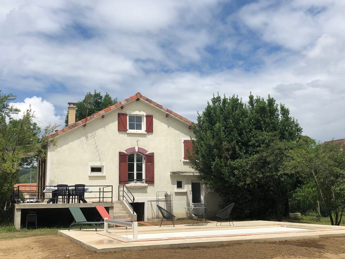 The exterior of the house features a light-colored facade with red shutters. A terrace equipped with outdoor seating is visible, alongside a lawn area and surrounding greenery. The sky above showcases scattered clouds, creating a bright atmosphere.