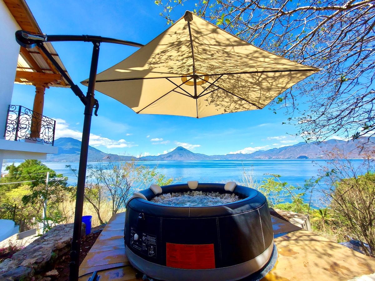 An outdoor hot tub is set on a wooden deck, shaded by a large umbrella. The scene showcases a sweeping view of the lake and surrounding mountains, with clear blue skies enhancing the tranquil setting.