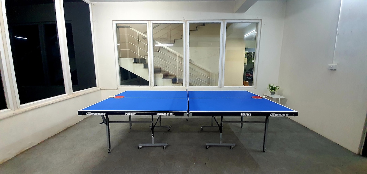 A spacious common area features two blue ping pong tables, providing recreational options for guests. Large windows allow ample natural light to fill the room. A staircase is visible in the background, ensuring easy access to other levels of the property.