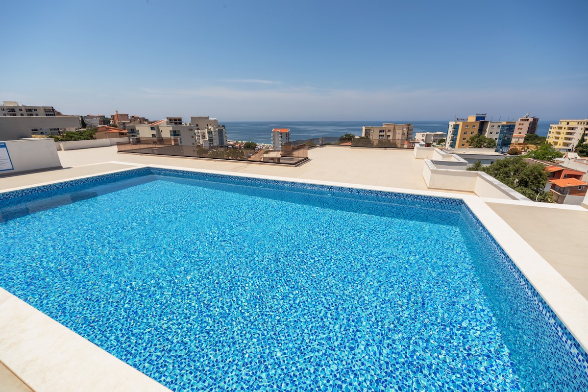 An outdoor pool features clear blue water, reflecting the bright sky. The pool area overlooks the sea, offering unobstructed views of the horizon and nearby buildings. The surrounding deck is spacious, providing ample room for relaxation and sunbathing.
