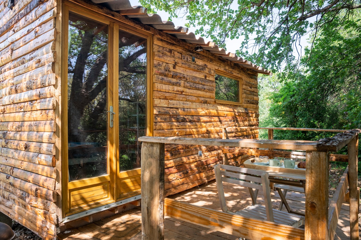A wooden chalet is shown, featuring a welcoming exterior with a covered terrace. Large glass doors offer a view of the surrounding greenery, while a simple table and chairs invite relaxation. The natural wood finish complements the outdoor setting.