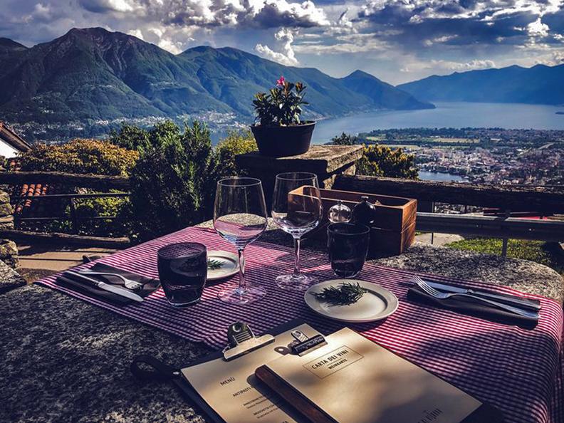 A dining table is set outdoors with a scenic view of mountains and a lake in the background. Glasses and dishes are arranged on a checkered tablecloth, complemented by a potted plant. A menu is placed on the table, enhancing the inviting dining space.