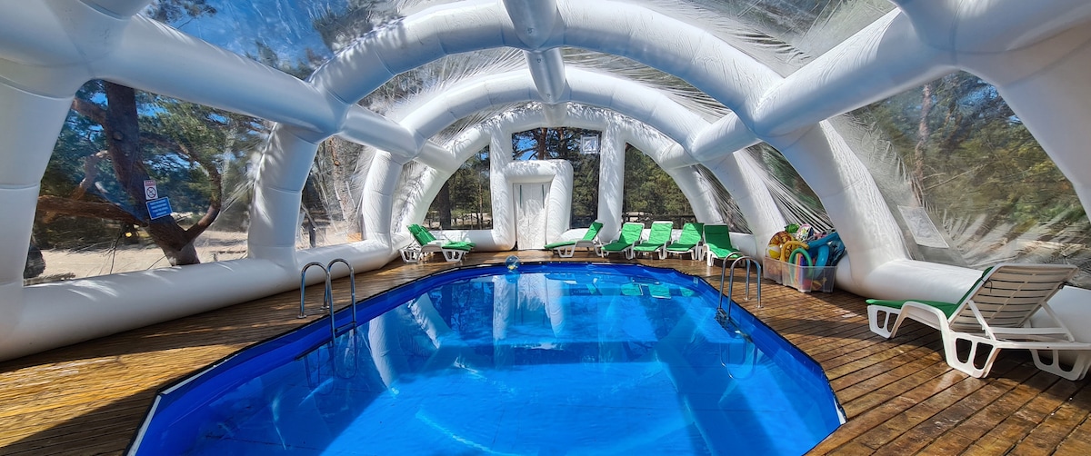 An indoor pool area is enclosed within a transparent inflatable structure. The pool is filled with clear water, surrounded by lounge chairs and sun loungers, offering ample space for relaxation. Sunlight filters in through the clear panels, providing a bright, welcoming atmosphere.