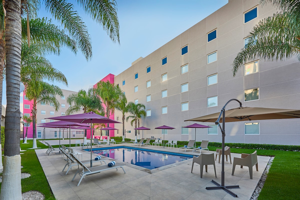 An outdoor pool area features several lounge chairs shaded by large umbrellas. Tall palm trees line the space, providing a tropical feel. The building's exterior is light-colored, with multiple windows reflecting the sky, creating a bright and inviting atmosphere.