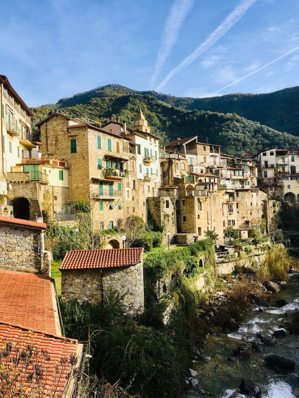 A scenic view of a medieval village built on a hillside. Stone and timber buildings with colorful facades are nestled against the mountain, overlooking a flowing river. Lush greenery surrounds the structures, complementing the natural landscape under a clear blue sky.