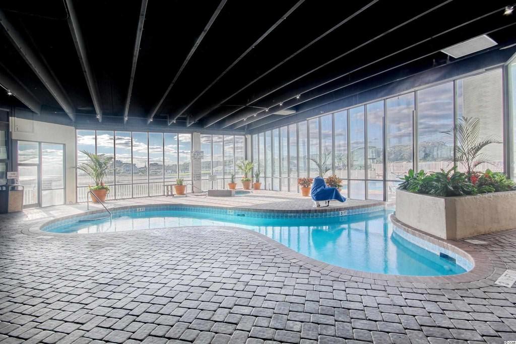 An indoor pool area is surrounded by expansive glass windows, allowing natural light to fill the space. Potted plants are placed strategically around the pool, which features a curvy design. The slate-tiled flooring enhances the modern feel of the environment.