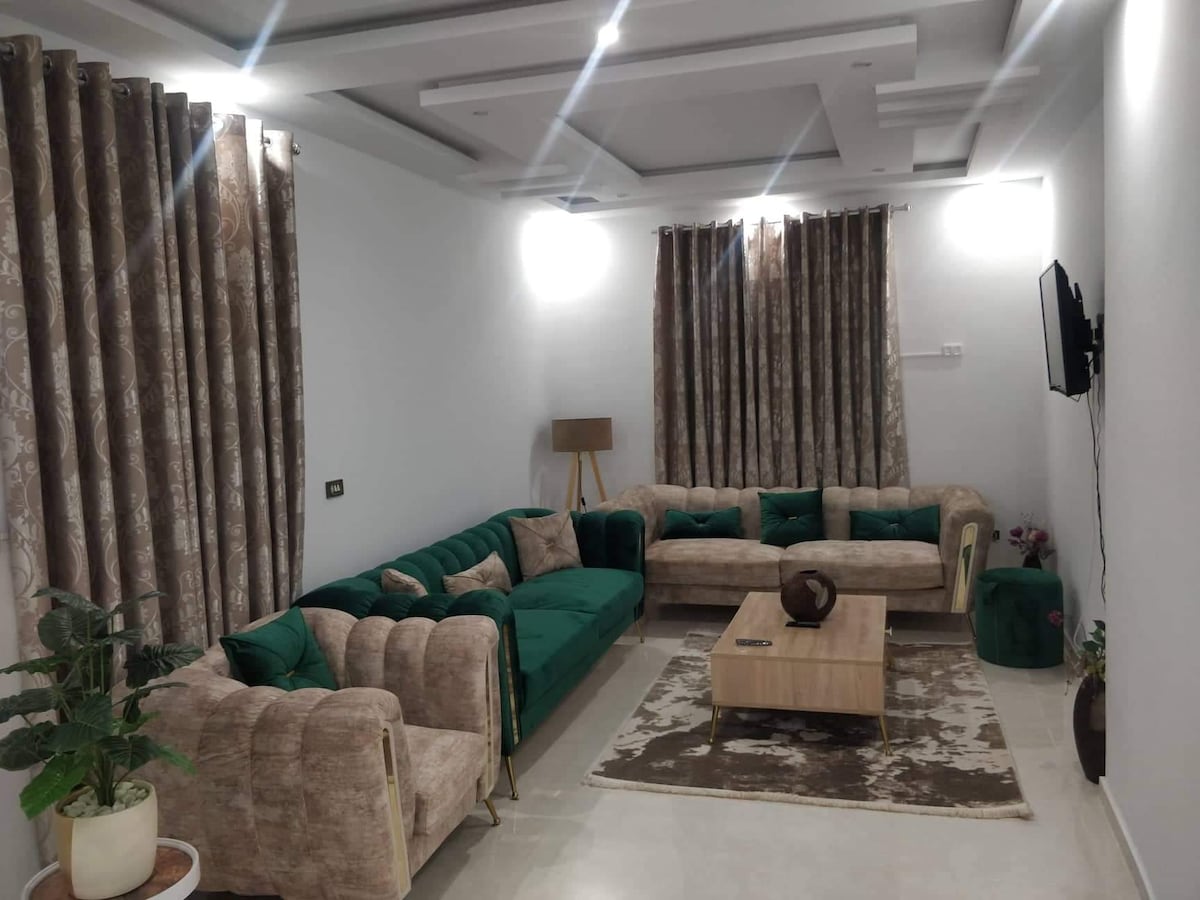 A spacious living area features a combination of green and beige seating arrangements, complemented by a wooden coffee table. Elegant curtains frame the windows, while a decorative plant adds a touch of greenery to the room's modern aesthetic.