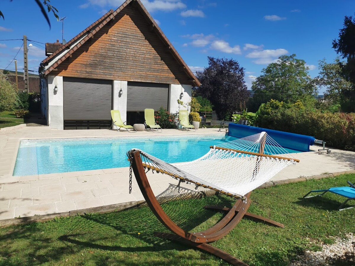 A wooden hammock is positioned on the lawn beside a refreshing pool. Sun loungers are arranged along the poolside, while lush greenery surrounds the space. A stylish house with a sloped roof is visible in the background, contributing to the serene outdoor environment.