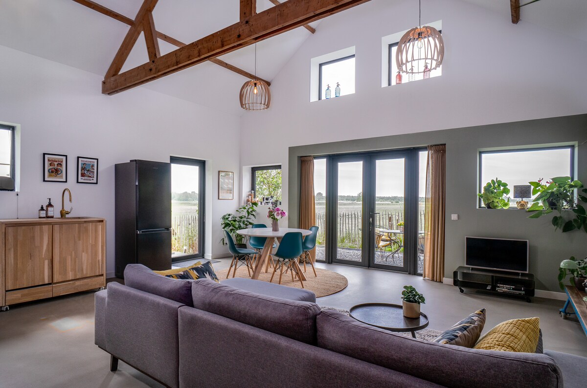 A modern living space features high ceilings with wooden beams. Large windows allow natural light, open to a scenic view. Seating includes a gray sofa and a dining table surrounded by colorful chairs. A kitchen area with wood cabinetry is visible, alongside a black refrigerator.