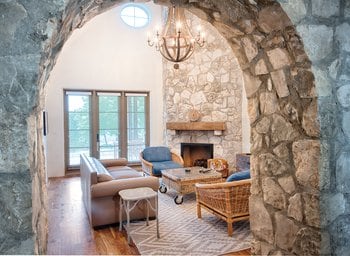 An archway leads into a spacious living area featuring a stone fireplace and vaulted ceiling. Large windows allow natural light, highlighting a cozy seating arrangement with a mix of upholstered and wicker furniture. A rustic coffee table is centered on a patterned rug.