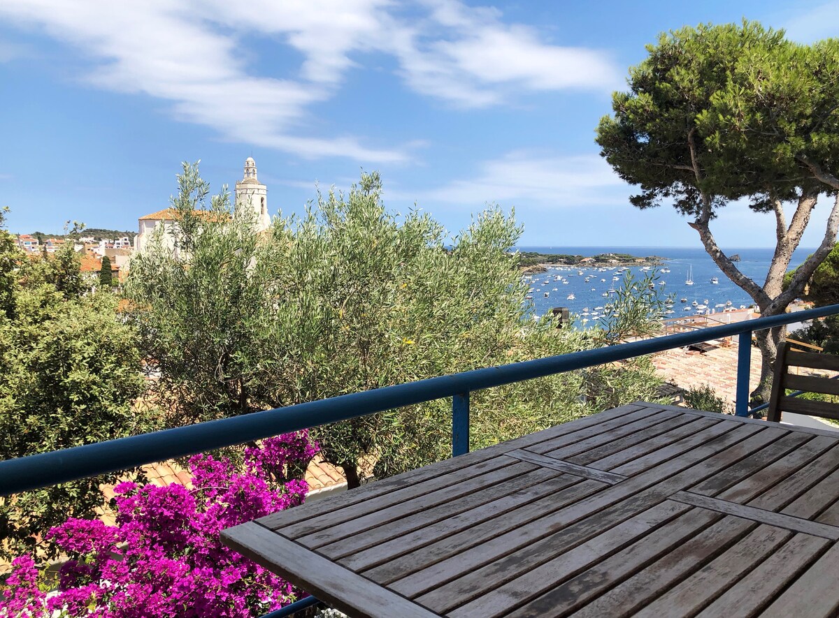A balcony features a wooden table with a view of the harbor and the sea. Bougainvillea in shades of pink adds a vibrant touch, while olive and pine trees provide greenery. The picturesque skyline includes a church tower and scattered boats on the water.