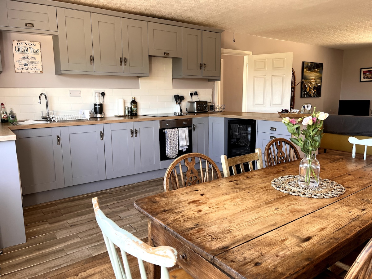 An open-plan kitchen and dining area features muted grey cabinetry and a wooden dining table surrounded by rustic chairs. A vase of flowers sits at the center of the table, while the kitchen is equipped with modern appliances, including a kettle and a coffee maker.