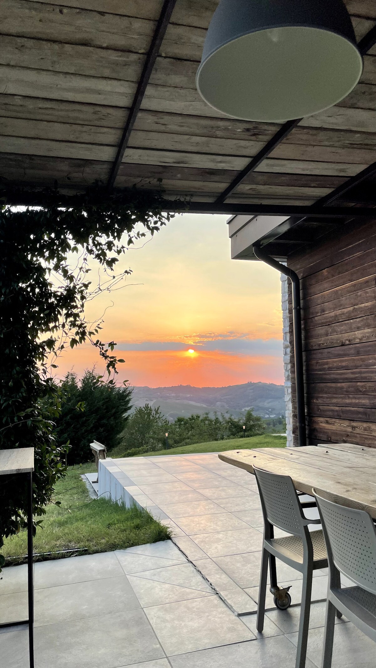 A stunning sunset is visible through the open space, framed by wooden and stone architecture. Outdoors, a spacious patio features a dining table with seating, inviting moments of relaxation while enjoying the natural beauty of the hills.