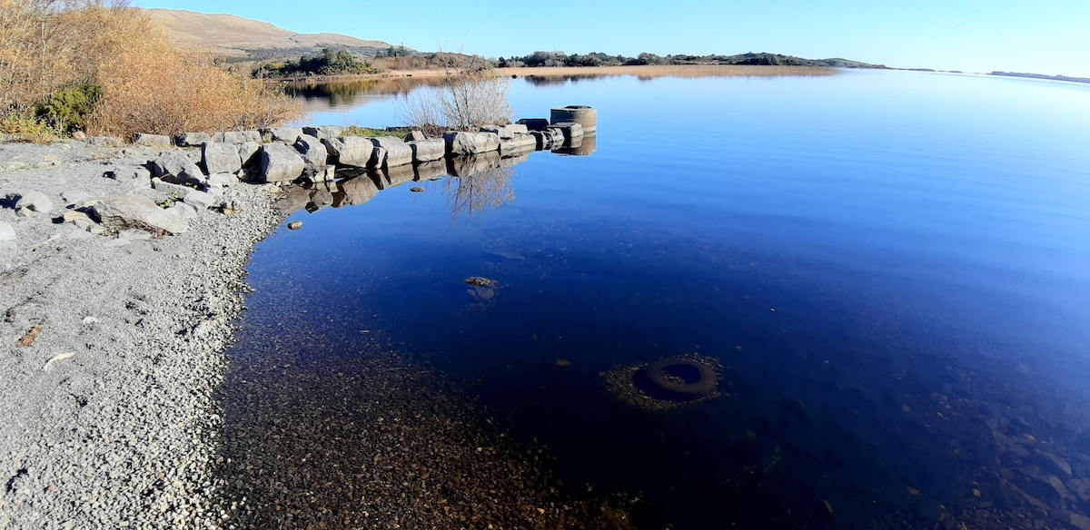 A serene lakeshore scene captures clear blue waters reflecting the sky, with a rocky jetty extending into the lake. Nearby, sparse vegetation and smooth pebbles line the water’s edge, creating a tranquil atmosphere.