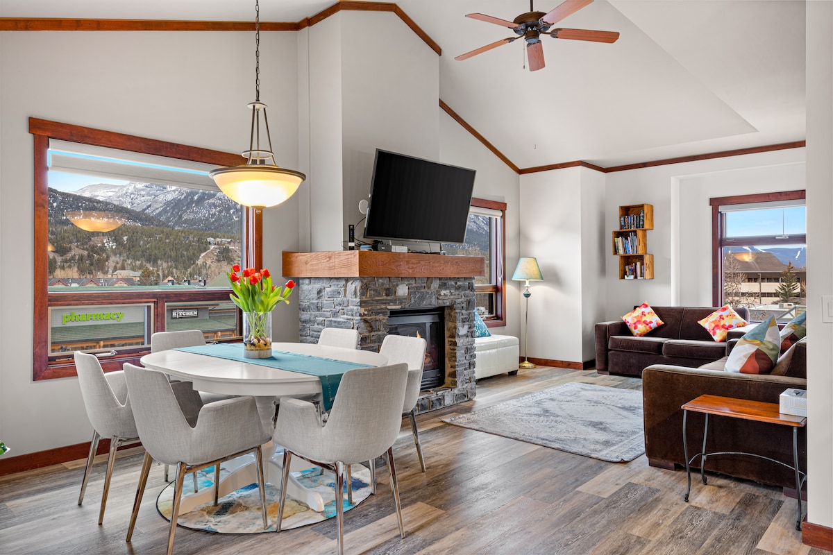 A bright living area features a spacious dining table surrounded by modern chairs, complemented by a cozy seating arrangement. Large windows reveal mountain views, while a gas fireplace offers warmth. A flat-screen TV is mounted above the stone hearth, adding to the inviting atmosphere.