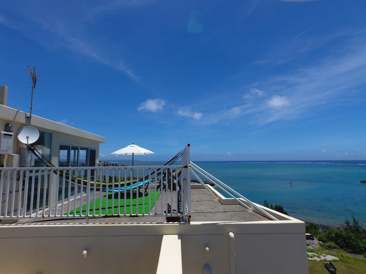 An expansive rooftop terrace features a sun umbrella and a clear view of the ocean. The blue sky complements the tranquil sea, with green grass covering part of the outdoor area. The surrounding landscape displays a combination of coastal and grassy terrain.