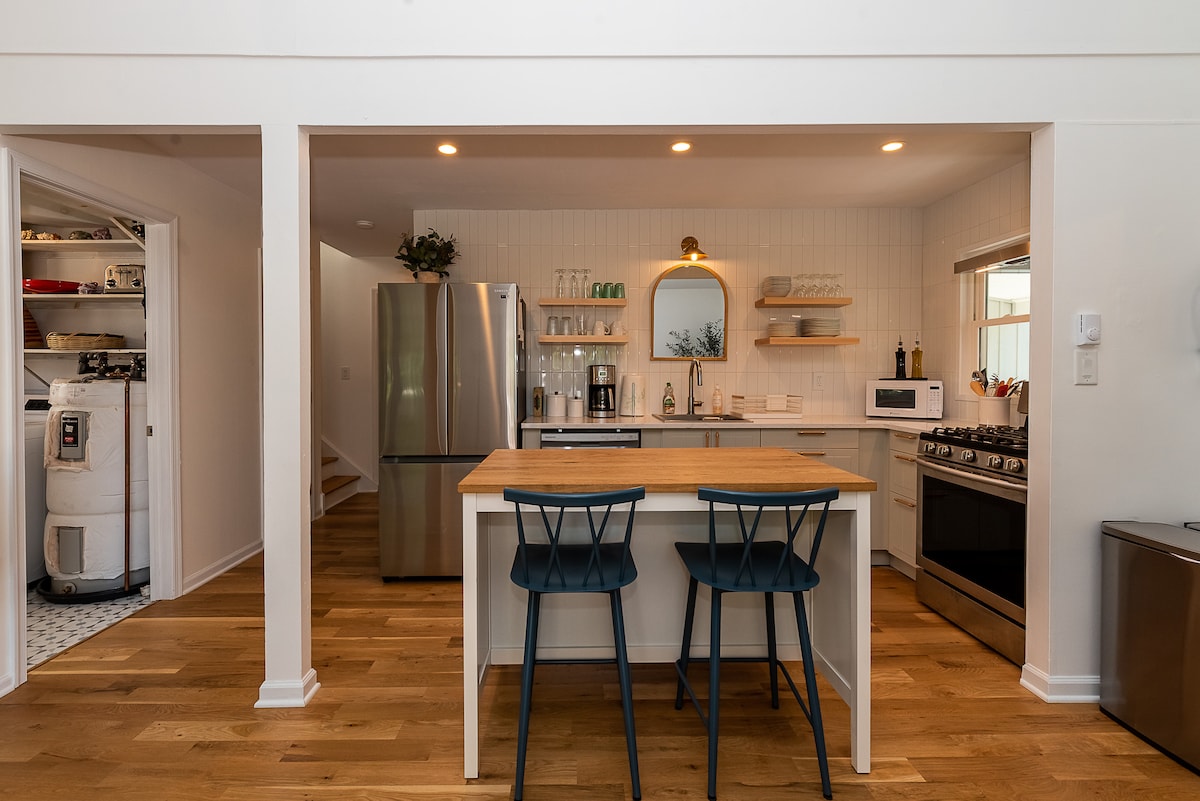 A modern kitchen layout features stainless steel appliances, including a refrigerator and gas range. A central island with two chairs provides space for casual dining. Open shelves host glassware, while a window allows natural light to brighten the space.
