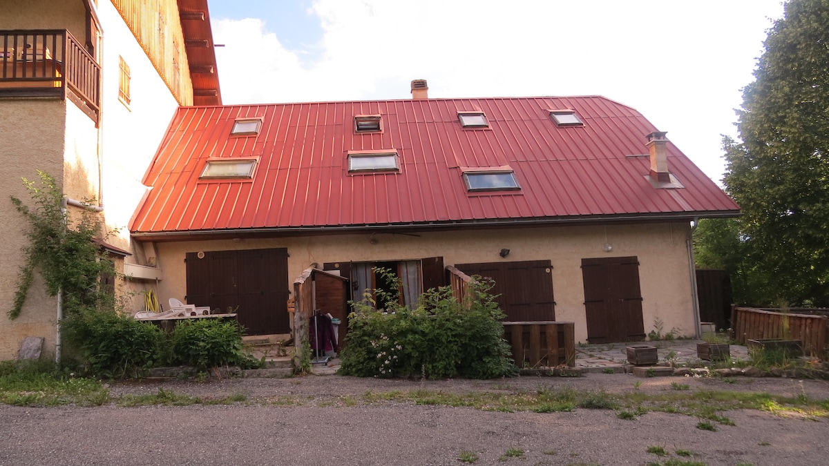 The exterior view of the residence showcases a distinctive red metal roof and wooden shutters. Lush greenery surrounds the entrance, where a small seating area is visible. The building's rustic charm is emphasized by the natural setting and the nearby stone patio.
