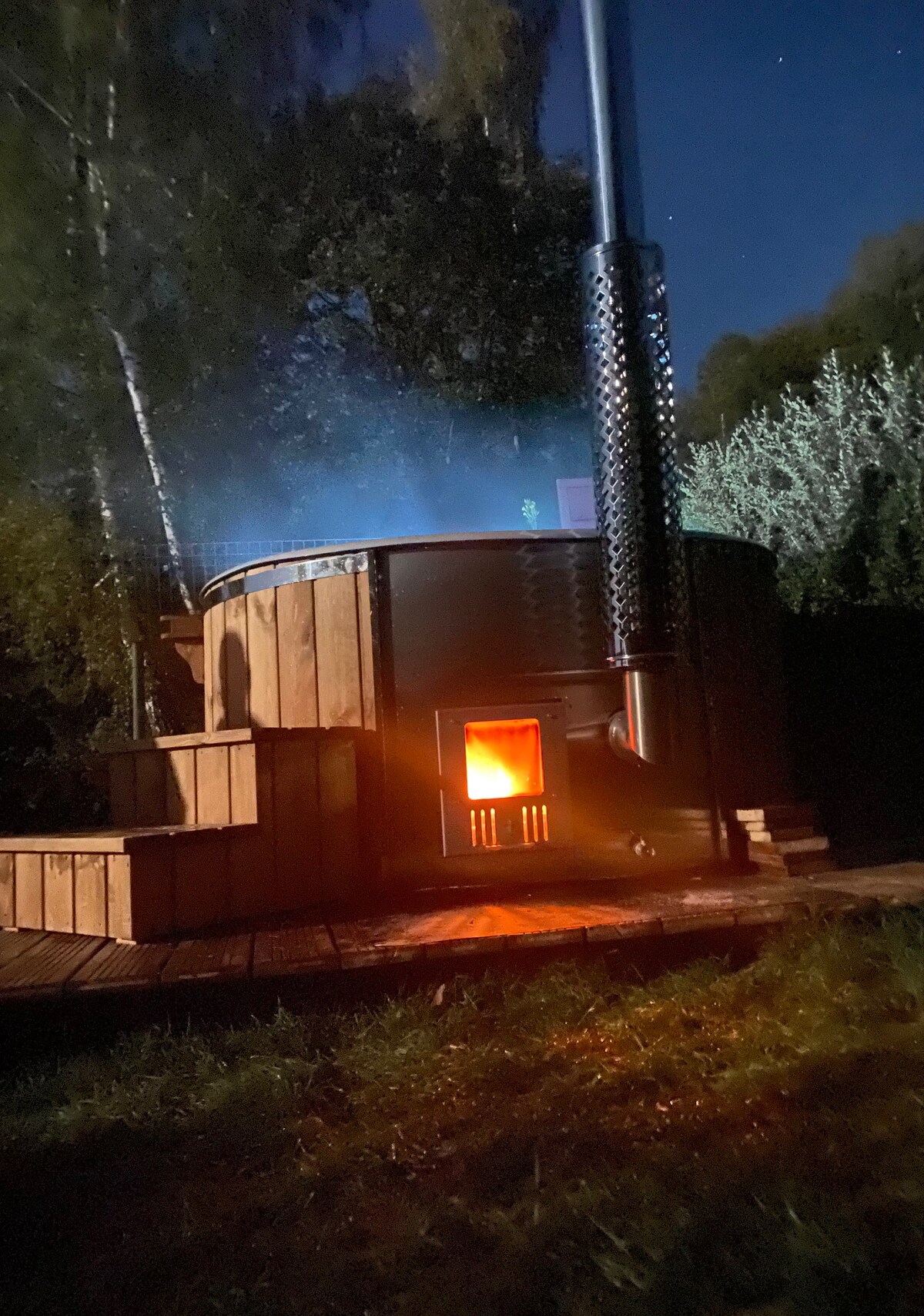 A wooden hot tub is illuminated by a warm glow from the fire inside, surrounded by stairs leading up to the tub. Soft steam rises into the night air, with trees visible in the background, creating a calming atmosphere.